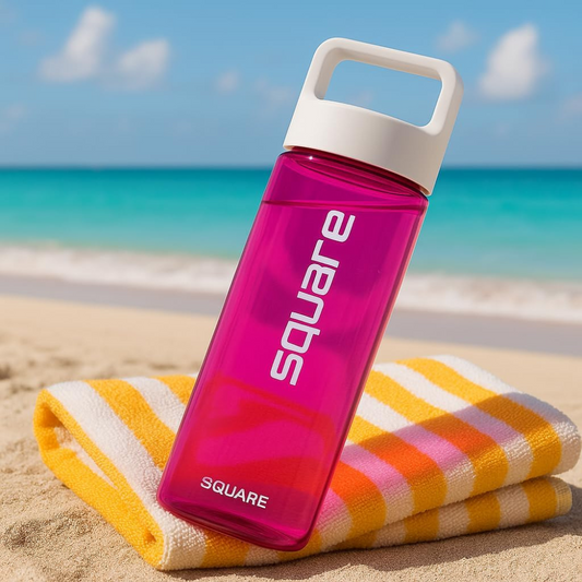 Bright pink reusable square water bottle with a white lid placed on a yellow-and-white striped towel at the beach, with turquoise ocean waves and blue sky in the background.